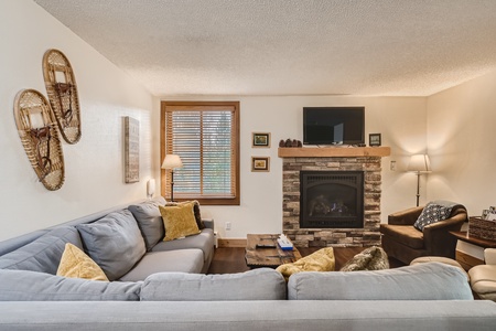 A cozy living room with a gray sectional sofa, yellow accent pillows, a stone fireplace with a TV above it, and a window with wooden blinds. Decor includes snowshoes and framed artwork on the walls.