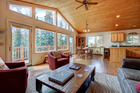 A spacious living area with a vaulted wooden ceiling, large windows showcasing a forest view, wooden furniture, a coffee table, a dining set, and a kitchen with wooden cabinets and a center island.