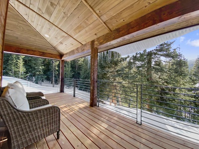 Covered wooden deck with two wicker chairs, overlooking snowy pine trees and an outdoor view of a winter landscape. The deck has a metal railing and is sheltered by a wooden ceiling.