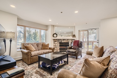 A cozy living room with two sofas, an armchair, a coffee table, and a fireplace. The room has large windows with blinds and a mix of decor, including a wreath above the fireplace.