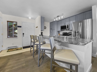 A modern kitchen with gray cabinets, marble countertops, and stainless steel appliances. A breakfast bar with four chairs lines the counter. A doorway and a window are visible in the background.