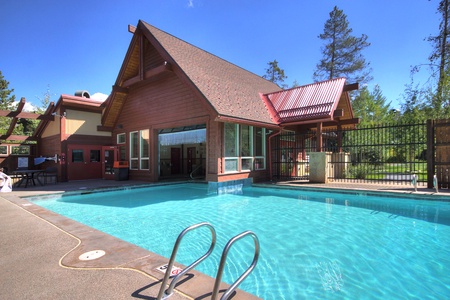 A modern outdoor swimming pool area with a wooden cabin-style building in the background, surrounded by trees, under a clear blue sky.