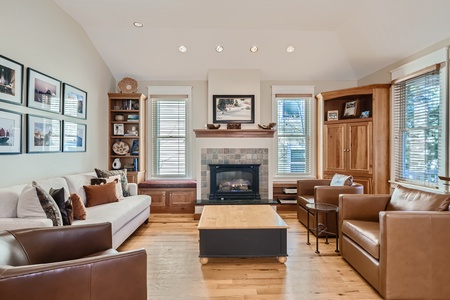 A cozy living room with a sofa, two armchairs, a wooden coffee table, and a fireplace. Large windows, framed pictures, and built-in shelves with decor items are also visible.