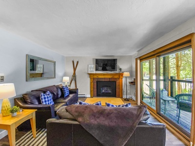 A cozy living room featuring a fireplace, leather sofas with plaid cushions, a wall-mounted TV, large mirror, and sliding glass doors leading to an outdoor patio with wicker chairs.