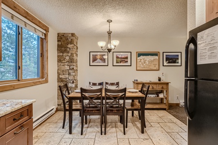 A dining area with a wooden table and six chairs, a chandelier above, wall art including pictures and a map, and a stone accent. The adjacent kitchen has granite counters and a black refrigerator.