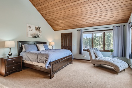 A bedroom with a sloped wooden ceiling, a large bed with blue bedding, two bedside tables with lamps, a wall painting above the bed, and a chaise lounge by a window with curtains.