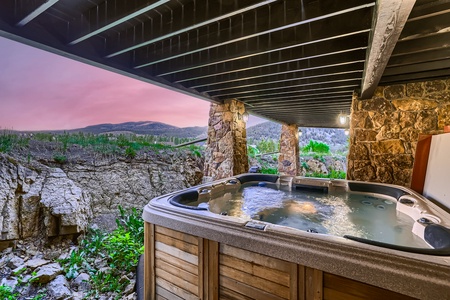 An outdoor hot tub with jets running, nestled under a wooded deck with stone pillars, overlooks a rocky landscape and distant hills during sunset.