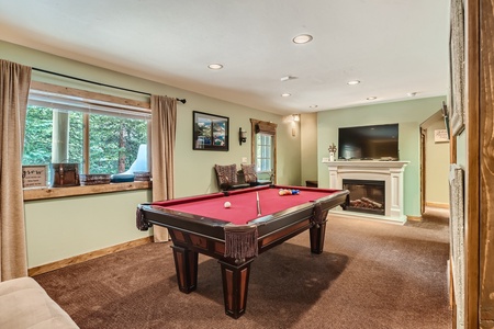 A basement room with a pool table, a fireplace with a TV above it, and a window with a green view. The room is furnished with a chair and decor items, creating a cozy entertainment space.