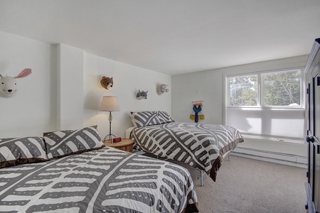 A bedroom with two double beds, each with black and white patterned bedding. There is a bedside table with a lamp and an alarm clock. Stuffed animal heads are mounted on the wall, and a window provides natural light.