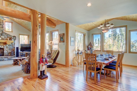 A spacious dining area with a wooden table and chairs, adjacent to a living room featuring a stone fireplace. Large windows offer views of a wooded landscape.