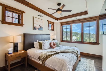 A well-lit bedroom with a large bed, wooden side tables with lamps, ceiling fan, and three windows offering a scenic outdoor view. The room features wooden trim and a cozy, inviting atmosphere.