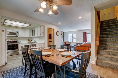 A dining area with a table set for six, adjacent to an open kitchen on the left. A carpeted staircase on the right leads to an upper floor. The living room with a sofa and a fireplace is visible in the background.