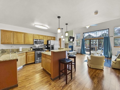 A spacious kitchen and living area with wooden flooring, modern appliances, granite countertops, and large windows providing a view of the outdoors.