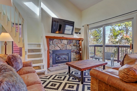 Living room with a stone fireplace below a mounted TV, a wooden coffee table, patterned rug, and sliding glass door leading to a balcony with outdoor chairs. Stairs are visible on the left.