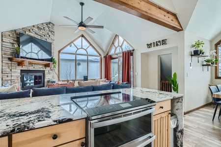 Spacious modern kitchen with a marble countertop, stainless steel stove, open shelving, and a view of a living area with a stone fireplace, large windows, and vaulted ceilings.