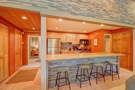 A modern kitchen with orange walls, stone accents, stainless steel appliances, and a wooden breakfast bar with four stools. A hallway with wooden doors is visible to the left.