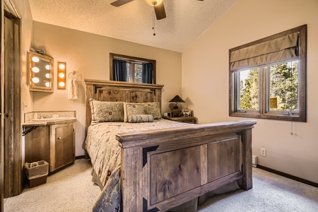 A bedroom with a wooden bed, two windows, a ceiling fan, a vanity with lights and a mirror, and beige walls.