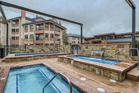 Outdoor spa area with two hot tubs surrounded by stone and metal structures, adjacent to a multi-story building on a cloudy day.