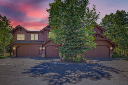 A two-story house with wood paneling and three garage doors, partially obscured by tall trees, set against a sky with a sunset glow.