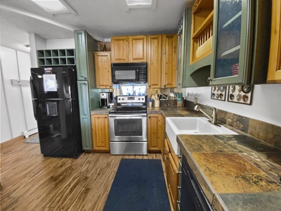 A kitchen with wooden cabinets, stainless steel appliances, a black refrigerator, a farmhouse sink, and a tiled countertop and backsplash. A blue mat is on the wooden floor.