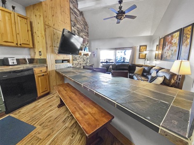 A cozy living area with a stone counter, wooden cabinets, a mounted TV, ceiling fan, sofa, decorative paintings, and a stone accent wall. Natural light enters through the window behind the sofa.