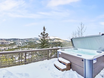 An outdoor hot tub with open lid sits on a snowy deck with a view of snow-covered mountains and trees in the background.