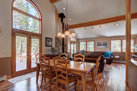 A rustic dining area with a wooden table and chairs adjacent to a living room with leather sofas. Large windows and a glass door provide a view of the trees outside.