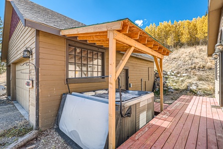 A covered hot tub with a wooden frame stands on a wooden deck attached to a beige wooden building with a window, set against a backdrop of trees and blue sky.