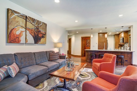 A modern living room featuring a gray sectional sofa, two orange armchairs, a wooden coffee table, and a painting of a deer above the couch. The kitchen area is visible in the background.