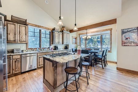 A spacious kitchen with wooden cabinets, a central island with barstools, pendant lighting, and a dining area in the background near large windows. A wall poster shows a Breckenridge ski theme.