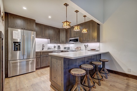 Modern kitchen with wooden cabinets, stainless steel refrigerator, built-in microwave, and three rustic barstools at a white countertop. Three pendant lights hang above the counter.