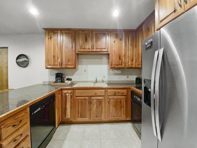 A kitchen with wooden cabinets, stainless steel refrigerator, tile flooring, and granite countertops. The sink is centered under upper cabinets, and kitchen appliances are on left and right counters.