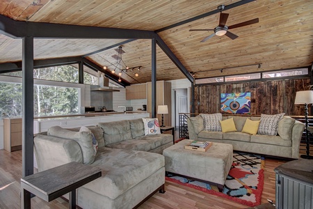 A spacious living room with wooden ceilings and large windows featuring a gray sectional sofa and a beige couch. The room includes a kitchen in the background, colorful decor, and a ceiling fan.