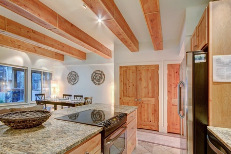 A modern kitchen with wooden beams and granite countertops. An oven, refrigerator, and dining area with a set table are visible. Two closed wooden doors are seen in the background.