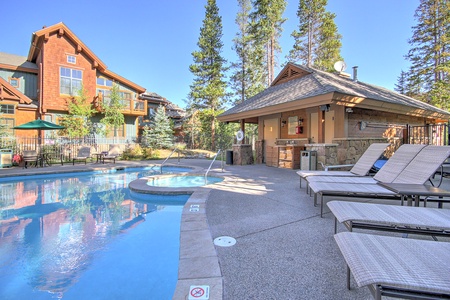 Outdoor pool with a hot tub surrounded by lounge chairs and a small shaded cabana area, set next to mountain-style buildings and tall trees.