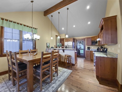 A kitchen and dining area with a wooden dining table and chairs, pendant lights over the kitchen island, granite countertops, wooden cabinetry, and a high ceiling with exposed beams.