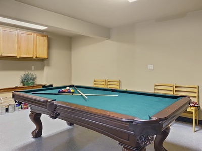 A room with a green felt pool table, pool cues, and a set of pool balls. Wooden chairs are against the wall, and wooden cabinets are mounted above a bench along the opposite wall.