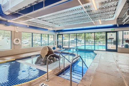 Indoor swimming pool with a waterslide, lounge chairs, and large windows showing views of outside greenery. The pool area is bright and spacious with overhead lighting.