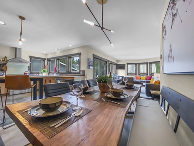 Modern open-concept dining and living room featuring a wooden dining table with place settings, bar stools at the kitchen counter, large windows, stylish light fixtures, and contemporary furnishings.