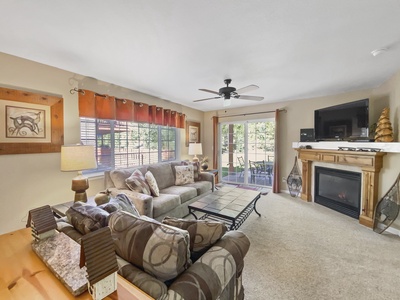 A well-lit living room with a large sofa, patterned pillows, a coffee table, a wall-mounted TV above a fireplace, and a ceiling fan. There are windows and a glass door opening to an outdoor area.