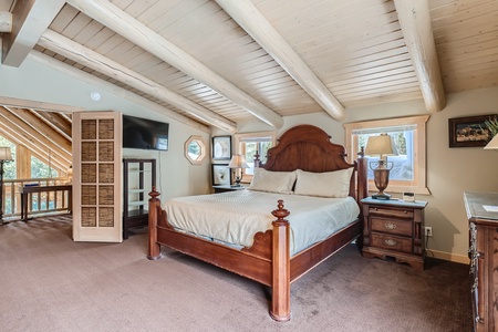 A bedroom with a wooden bed, two bedside tables with lamps, a television, and slanted wooden ceiling beams. There's a window and a half open door leading to another area.