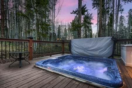 A hot tub, partially covered, sits on a wooden deck surrounded by a forested area. Steam rises from the water, and the sky shows a purple-pink sunset.