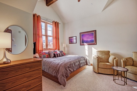 A cozy bedroom with a large bed, wooden dresser, wall-mounted mirror, two beige armchairs, a small round table, and a window with orange curtains. Two framed pictures hang on the wall.