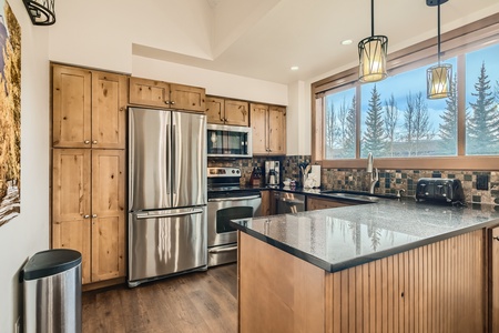 A modern kitchen with wooden cabinets, stainless steel appliances, a granite countertop island, two pendant lights, and a large window with a view of trees.