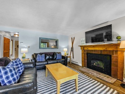 A living room with a striped rug, black leather couches with blue-checkered pillows, a wooden coffee table, a fireplace with a TV mounted above, and wall-mounted skis next to the fireplace.