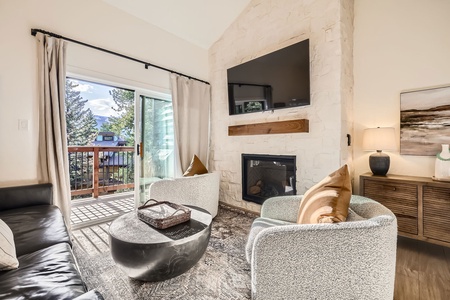 Cozy up in the stylish living room, featuring modern furnishings, a unique antler chandelier, and floor-to-ceiling windows that open to a private balcony with breathtaking forest and mountain views.