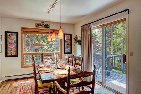 A dining room with a wooden table set for six, pendant lighting, and a sliding glass door leading to a deck with a grill. Artwork hangs on the walls, and a window offers a view of trees outside.