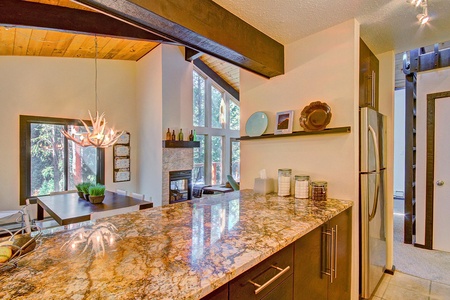 Modern kitchen and dining area with a marble countertop, stainless steel appliances, and large windows allowing natural light to enter. The dining table features a unique chandelier and potted plant.