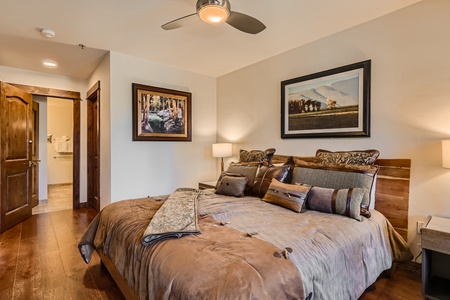 A bedroom with a large bed, decorative pillows, and a folded blanket. The room features wooden floors, a ceiling fan, two framed art pieces on the walls, and a doorway leading to a bathroom.