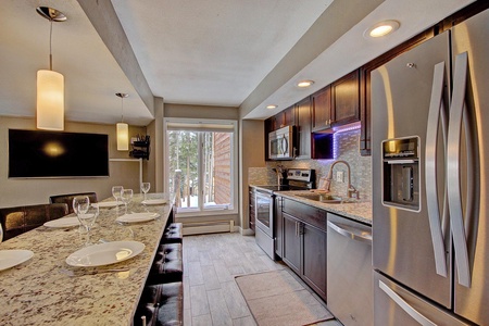 Modern kitchen with stainless steel appliances, granite countertops, and dark wood cabinets. A long island with bar stools and place settings is situated under pendant lighting. A large window illuminates the space.